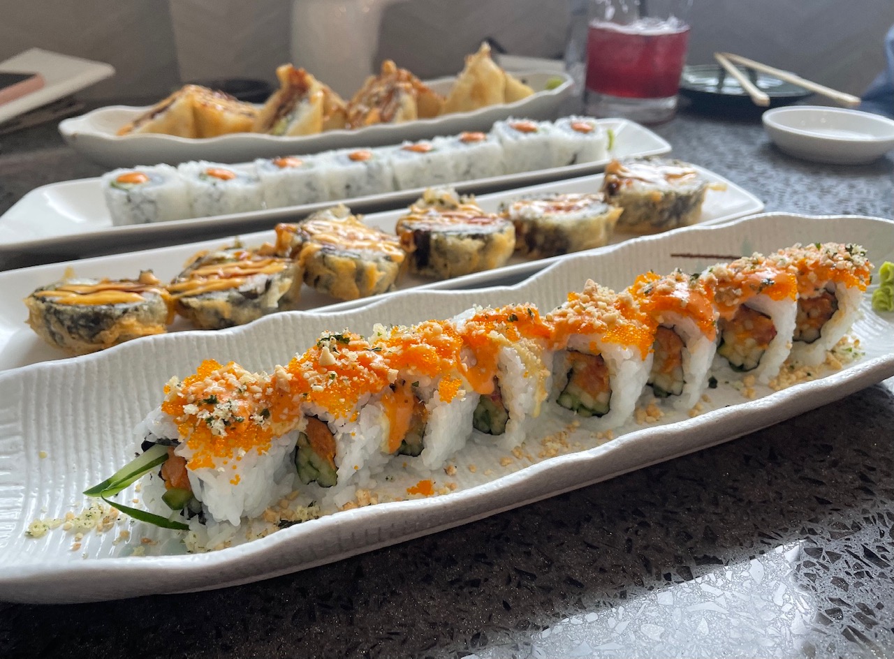 On a gray speckled table, there are four long white trays with a sushi roll on each. Photo by Alyssa Buckley.