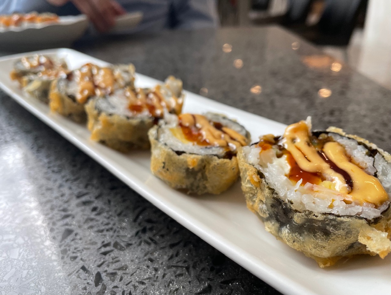 On a long white rectangular plate, there is a fried sushi roll with drizzles of sauce on it. Photo by Alyssa Buckley.