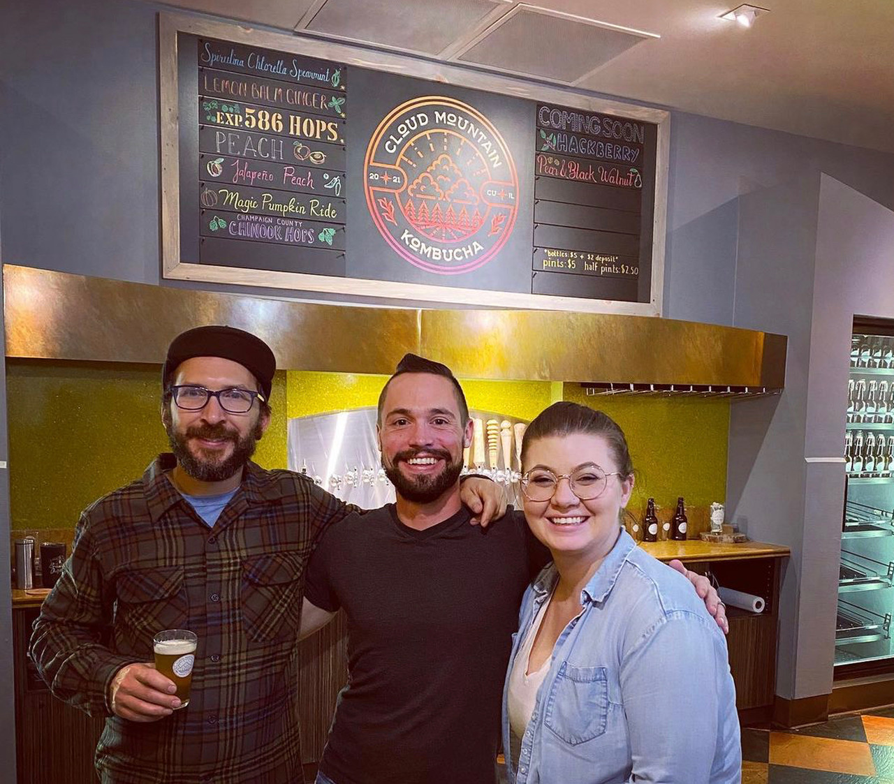 In front of the kombucha taps, there are three owners, all white. On the left is a bearded man in glasses and a hat holding a pint glass full of kombucha with his other arm around the bearded man in the middle who has his arm around a female. All three are smiling to the camera. Photo from Cloud Mountain Kombucha's Instagram page.