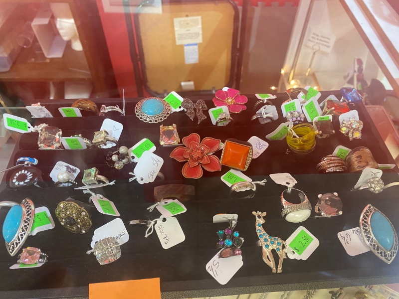 An assortment of rings are in a black display box within a glass case, most with a large ornamental piece. Each has a white price tag attached. Photo by Julie McClure.