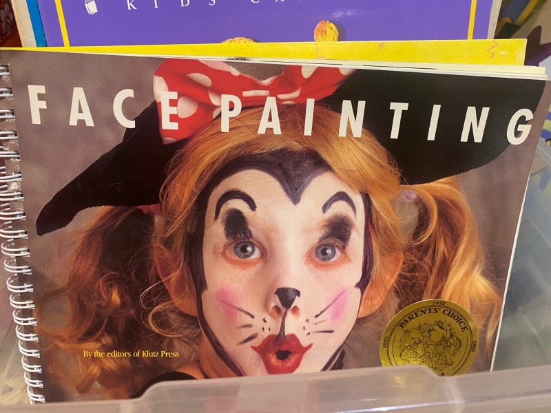 A book with a spiral binding. The cover shows a photo of a child in a wig with their face painted like a clown. It says Face Painting in white block letters across the top. Photo by Julie McClure.