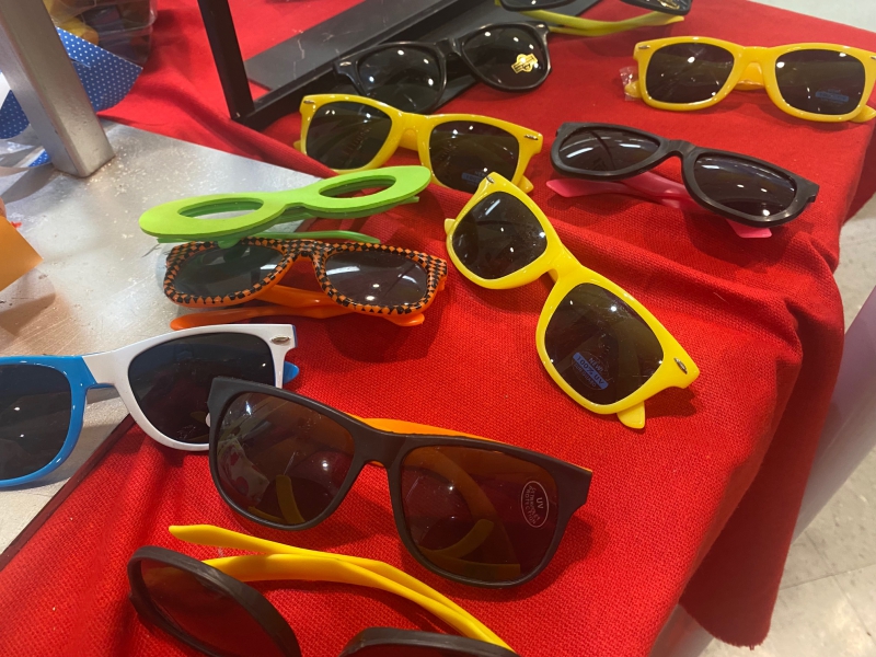 An assortment of sunglasses with solid colored frames are scattered on a table with a red drape. Photo by Julie McClure.