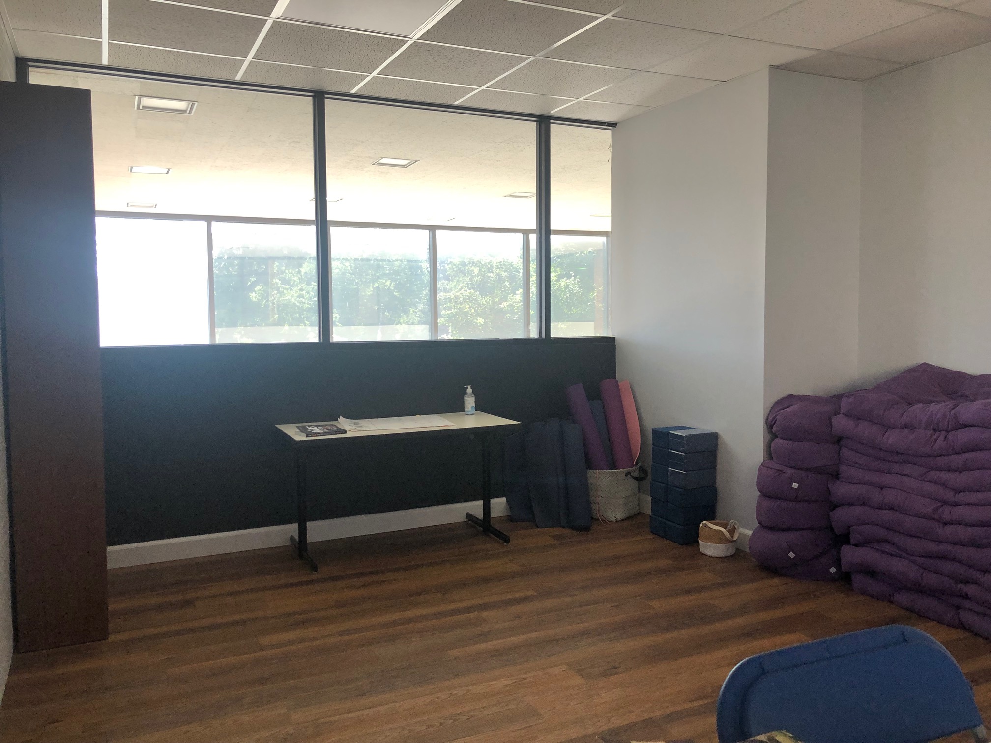 In the corner, there is a long wooden table with a single plastic water bottle on it. Along the right wall, there are stacked yoga mats and workout materials in front of a wood floor. Photo by Alyssa Buckley.
