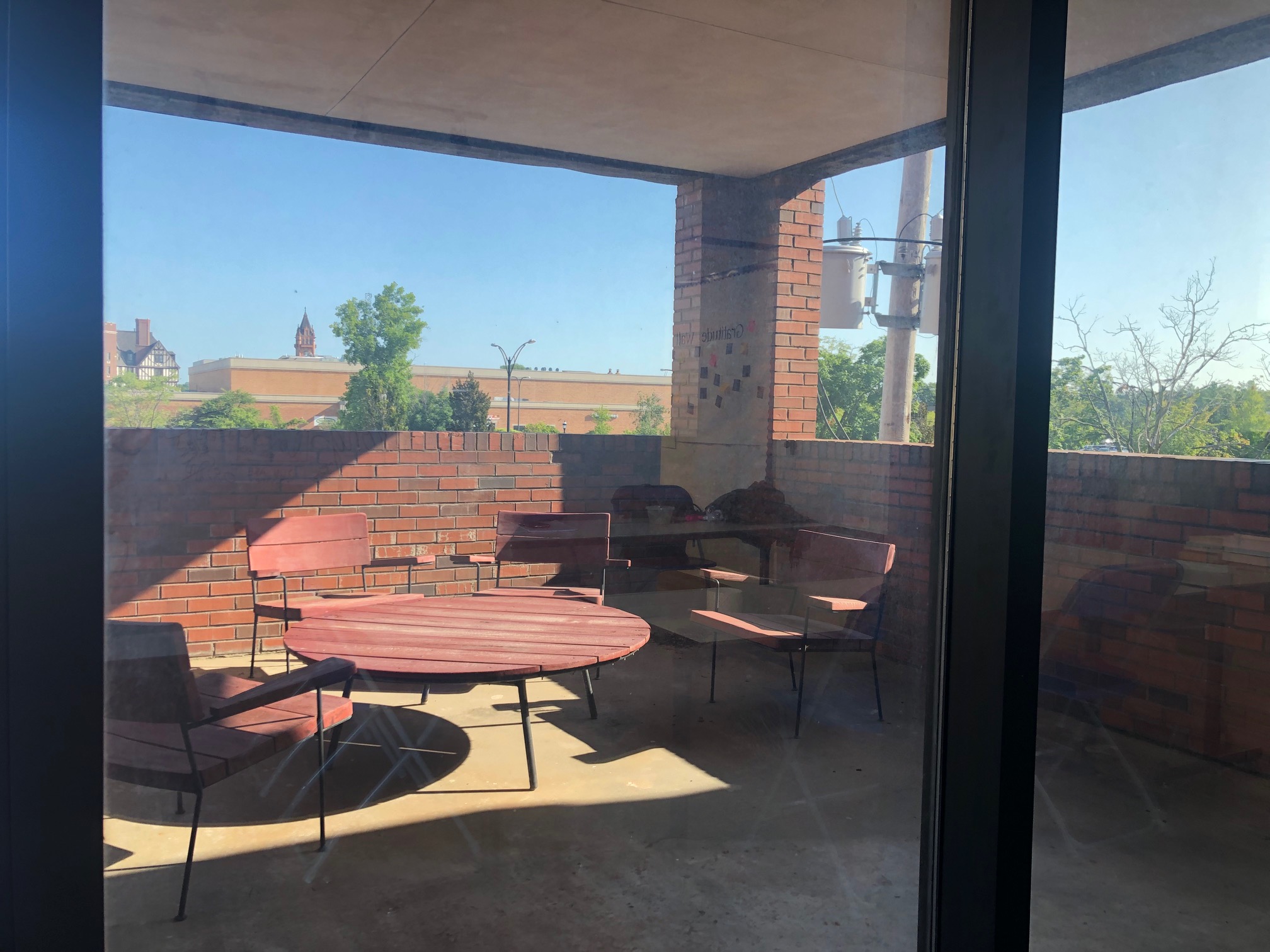 The outdoor space is seen through a window, showing a large circular coffee table and four large brown outdoor chairs with arms. Photo by Alyssa Buckley.