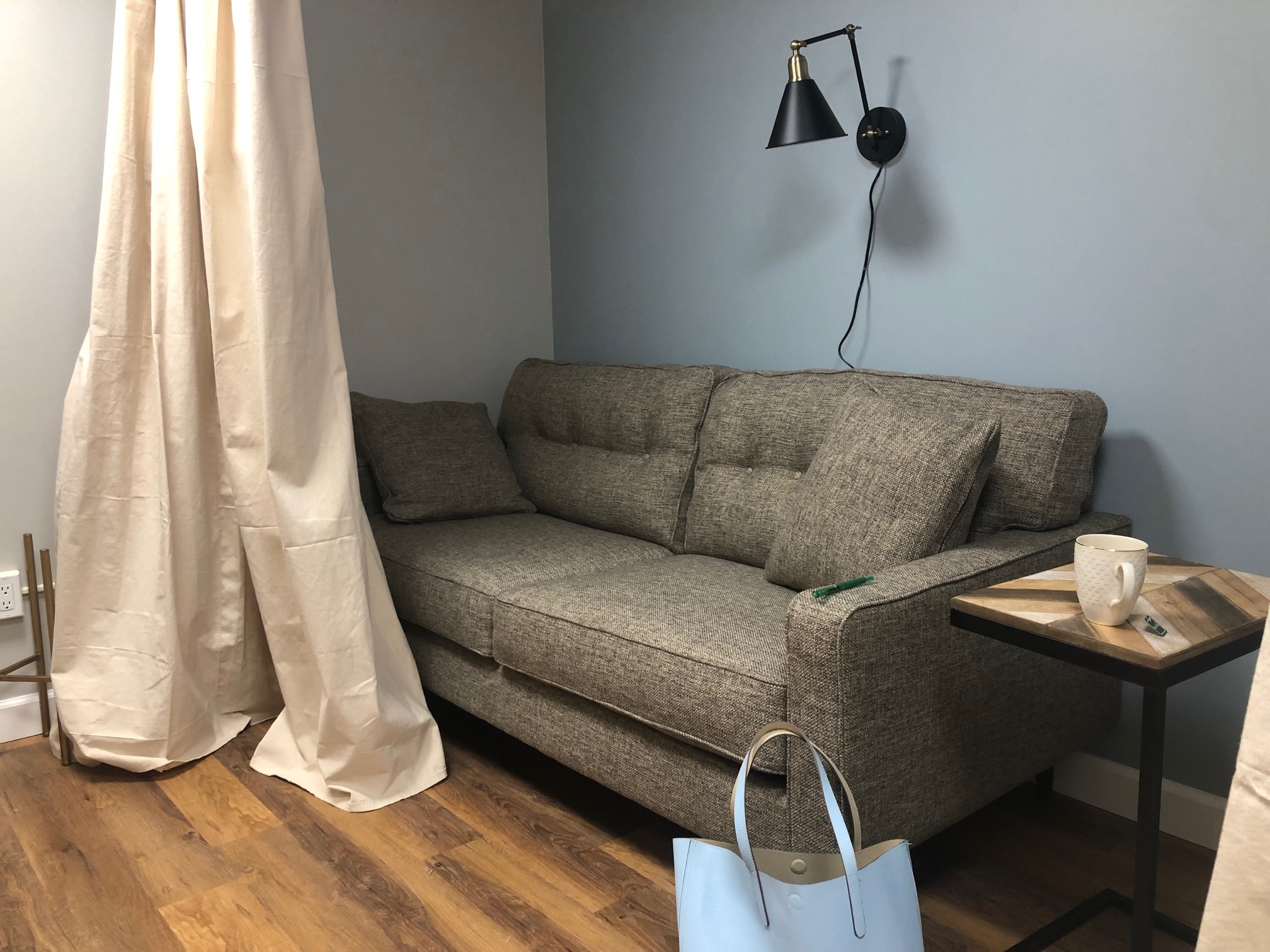 A large cushioned gray couch is in front of a gray wall with a curtain right in front of the couch. The wooden floor has a blue purse on it beside the couch, and there is an end table with notebooks and pens on it. Photo by Alyssa Buckley. 
