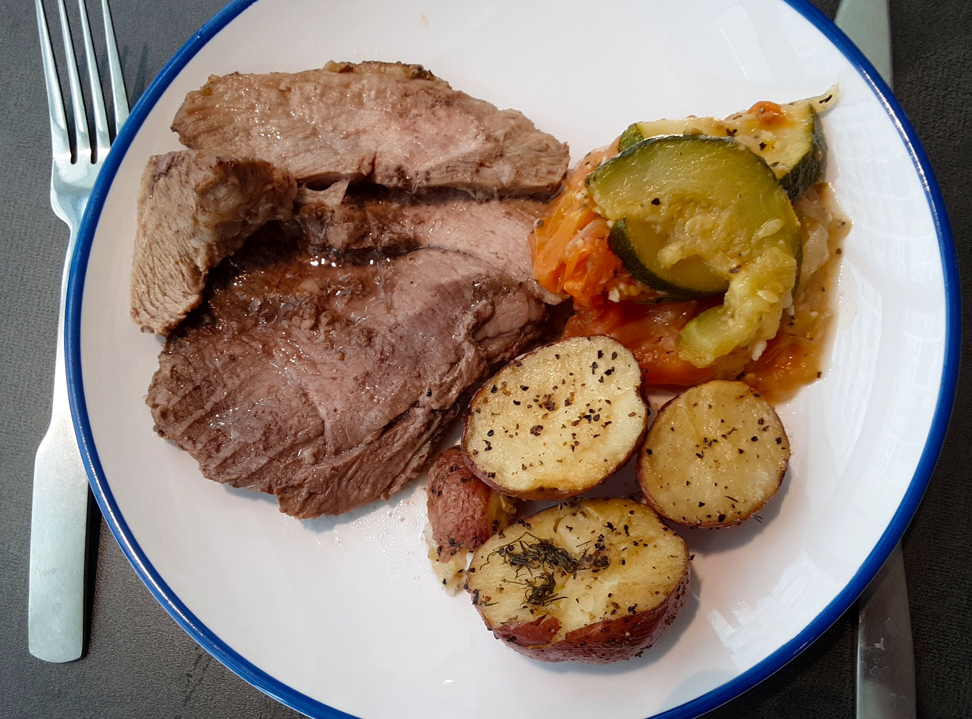 On a circle white plate with a blue rim, there are a few pieces of sliced lamb beside herb roasted potatoes and a side of mushy zucchini. Photo by Paul Young.