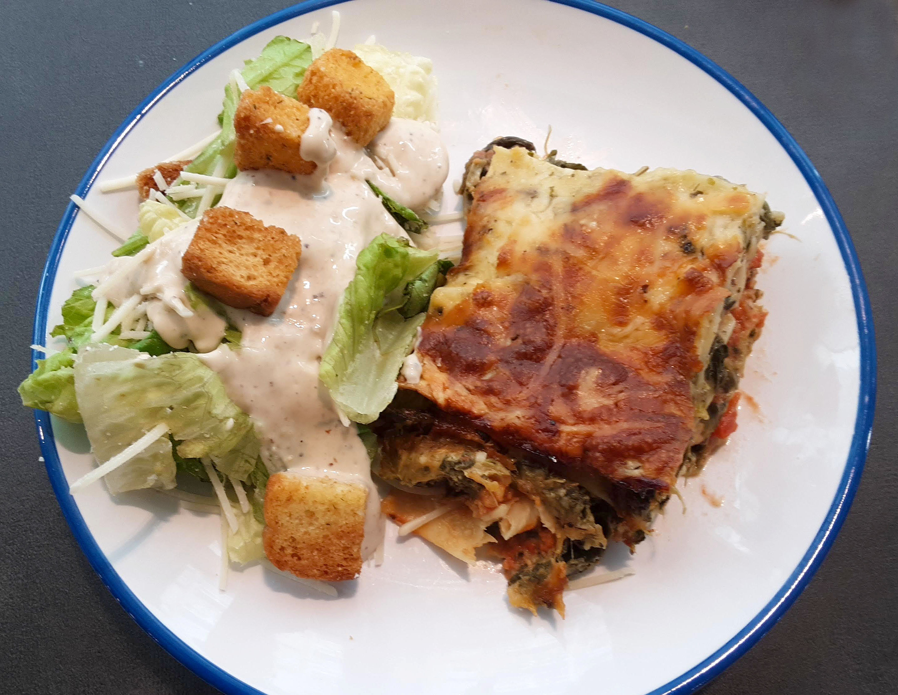 On a white circular plate with a blue rim, there is a lasagna with a side salad. Photo by Paul Young.