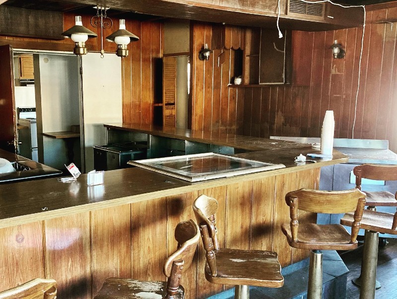 A portion of a bar area in an abandoned hotel. The walls are wood paneled, and the bar and bar stool are all wooden. A light fixture with two bulbs hangs above the bar. Photo from Champaign County Restore Facebook page.