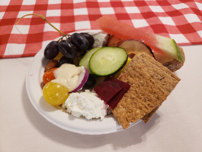 A small plate of food on a white tablecloth with a red checkered tablecloth in the background. On the plate there are crackers, bread cheese, tomatoes, cucumbers, grapes and watermelon. Photo by Sara Ressing.