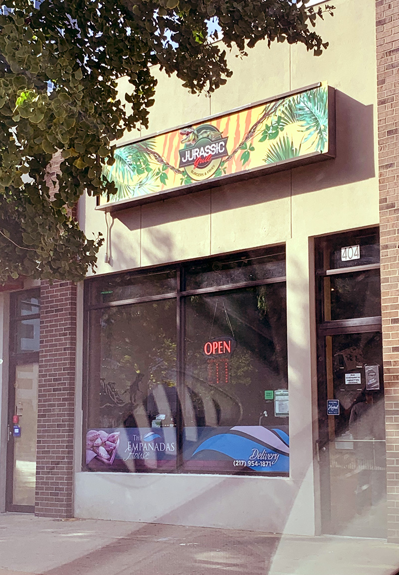 On Green Street in Campustown, the exterior of Jurassic Grill has a big window with an illuminated OPEN sign. Photo by Zoe Valentine. 