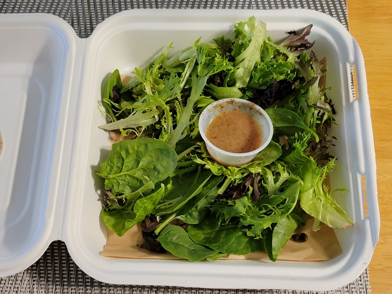 A large side salad styrofoam box with a cup of vinaigrette in the middle. Photo by Matthew Macomber.