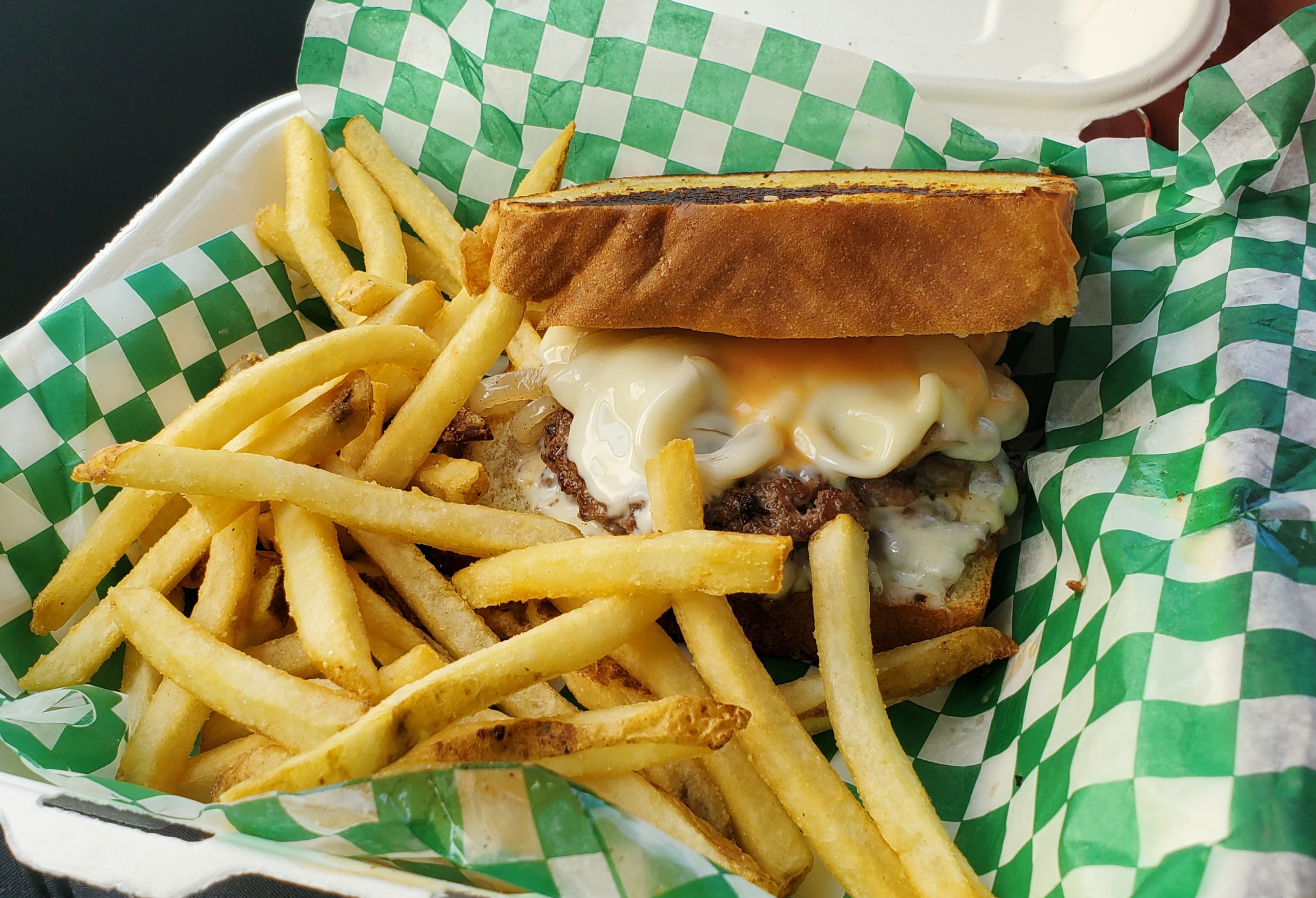 In a white styrofoam square takeout container lined with green and white checkered parchment paper, there is a burger with melted white cheese and a big portion of fries. Photo by Carl Busch.