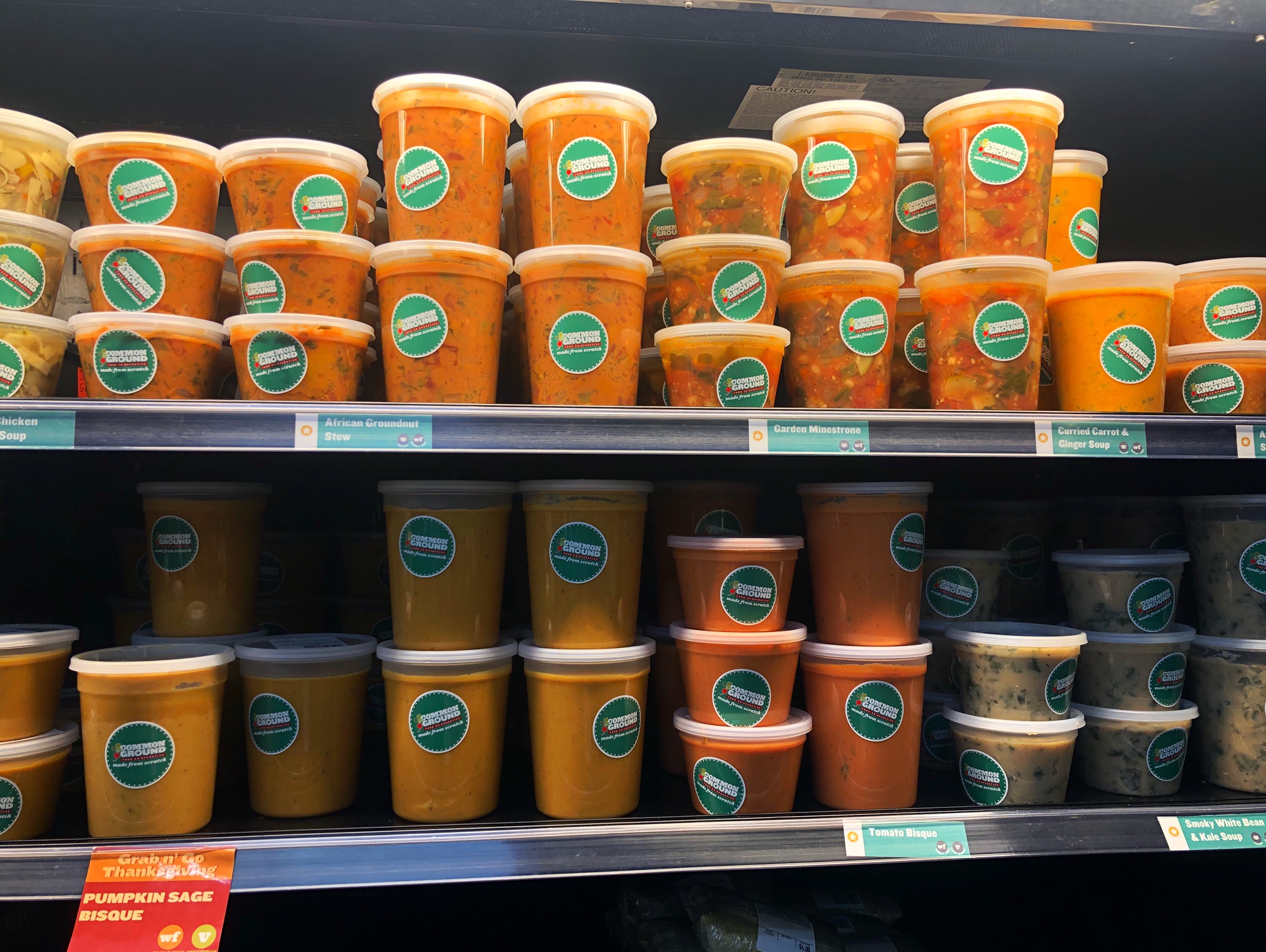 On two shelves, tall cylindrical plastic containers hold orange soups and sauces for sale at the grab-and-go section of Common Ground. Photo by Alyssa Buckley.