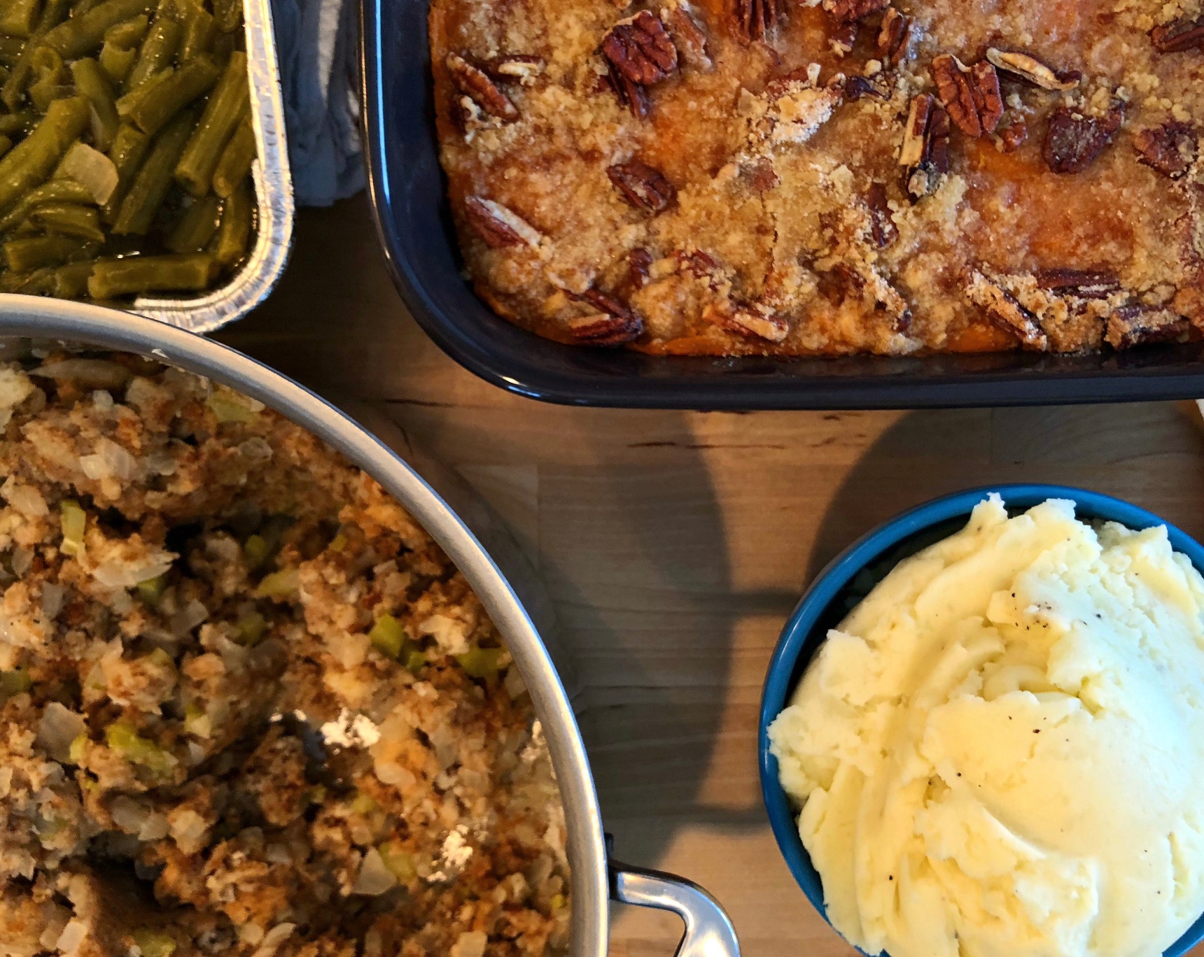 An overhead photo shows four side dishes in each corner. In the top left, green beans, top right, sweet potato casserole, bottom right, mashed potatoes in a blue bowl, and on the bottom right, stuffing in a metal pot. Photo by Alyssa Buckley.