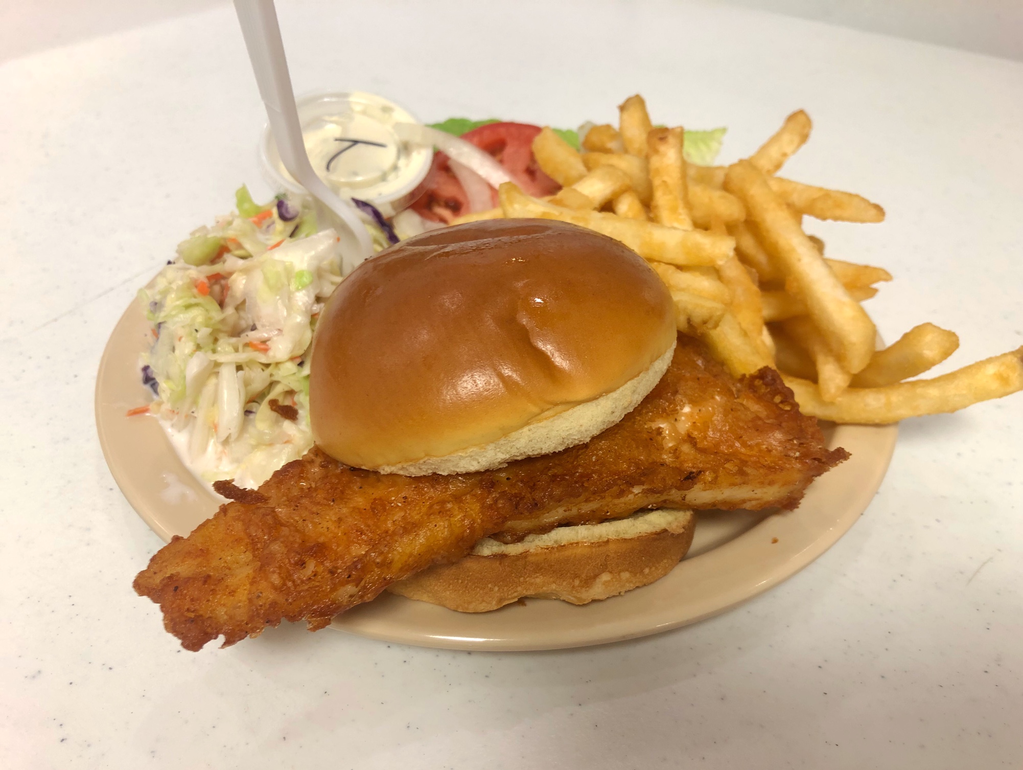 On a light brown plate, there is a fish sandwich with a filet of fish that sticks out both sides, a side of coleslaw with a plastic fork stuck in it, and a side of fries. Photo by Alyssa Buckley.