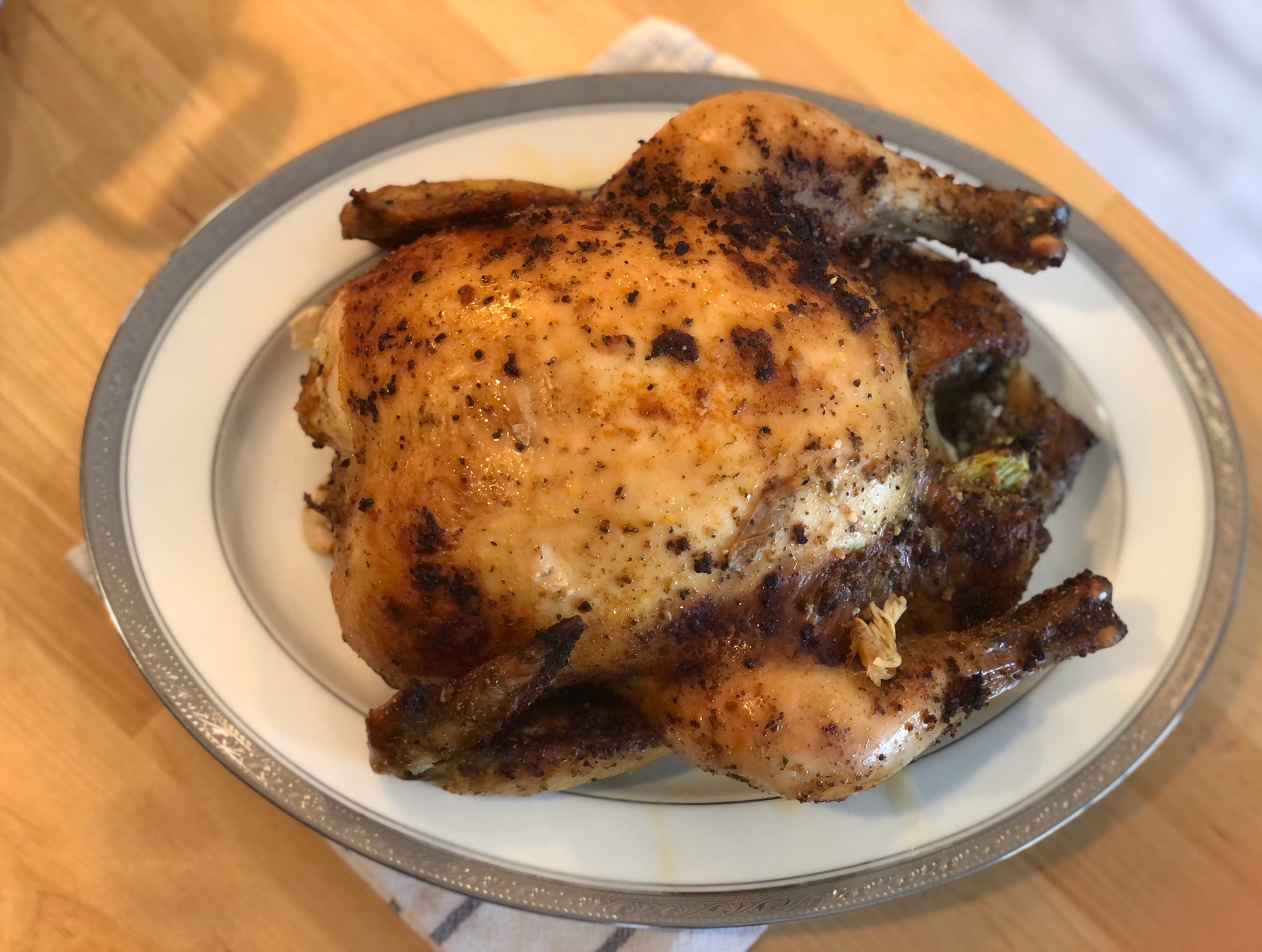 An overhead photo shows a fully cooked, seasoned whole chicken on a white platter with a silver border. Photo by Alyssa Buckley.