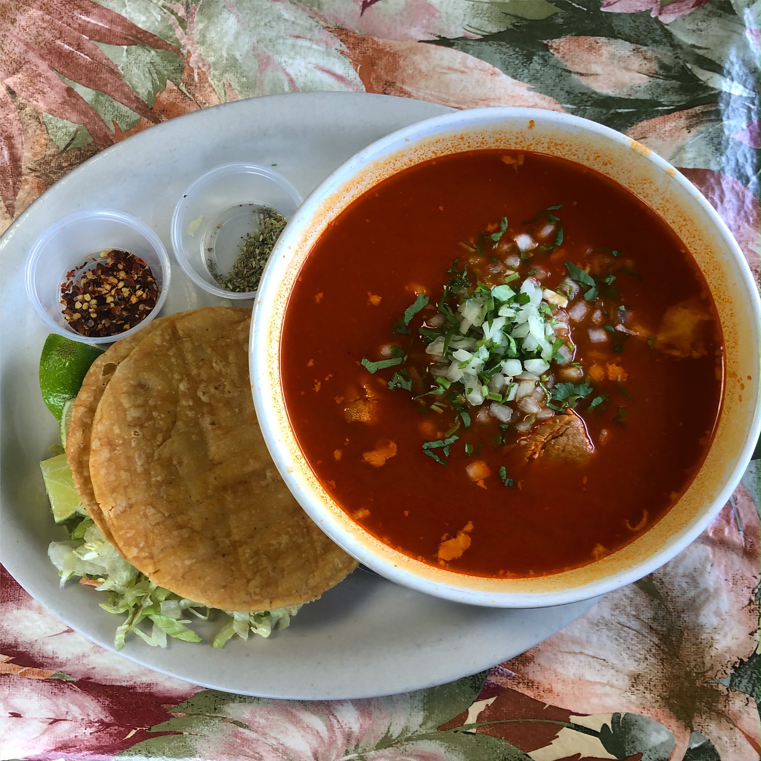 On a floral tablecloth, there is an oval plate with a large white bowl filled with red pozole soup. Beside the bowl on the oval plate, there are mini cups of dried seasoning, fried tostatas on lettuce, and lime wedges. Photo by Alyssa Buckley.