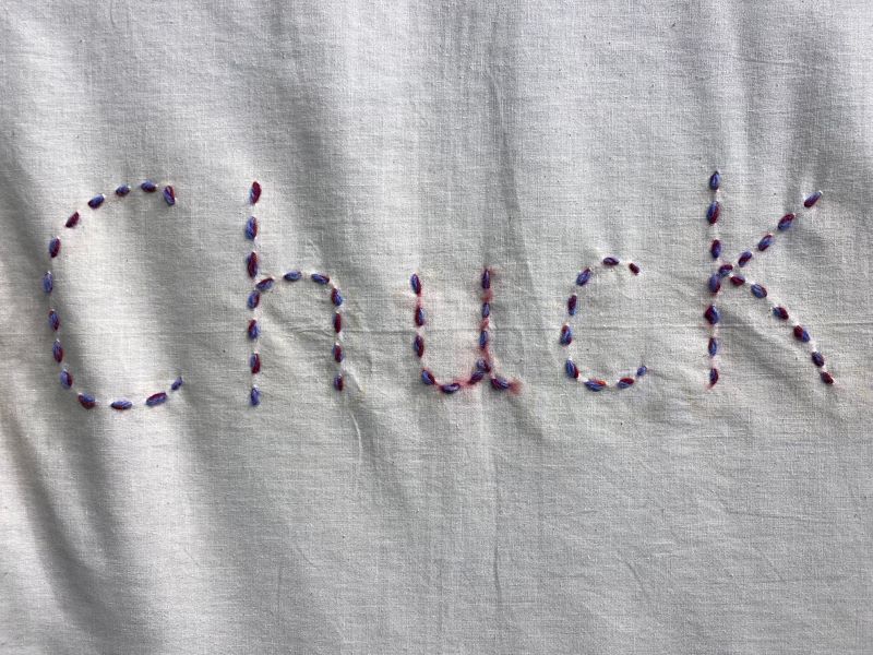 A close up of a quilt piece made of white linen. The name Chuck is stitched in blue and purple thread. Photo by Julie McClure with permission from Spurlock Museum.