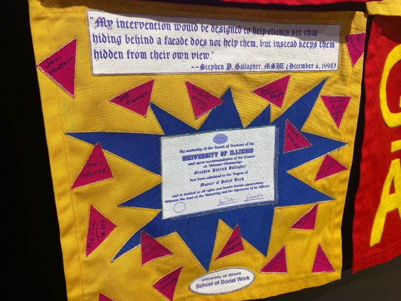 A close up of a quilt panel with a yellow background and starburst shape in blue. In the center of the shape is a copy of a U of I degree. There are red triangles with names scattered around the panel. Photo by Julie McClure with permission from Spurlock Museum.