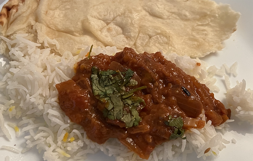 A close view of a spoon full of baingan bharta on top of rice with a torn piece of naan. Large slices of onion appear in a chunky red sauce of tomatos and eggplant. On the paingan bharta is a wad of green cilantro. Photo by Sara Ressing.