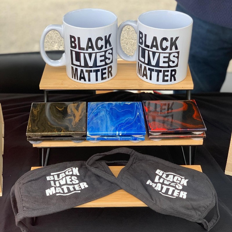 A three tiered wooden stand. The top shelf has white mugs that say Black Lives Matter in black lettering, the second shelf has multi colored marbled coasters, and the bottom shelf has masks with Black Lives Matter in white lettering. Photo from Buy Black Chambana Facebook page.