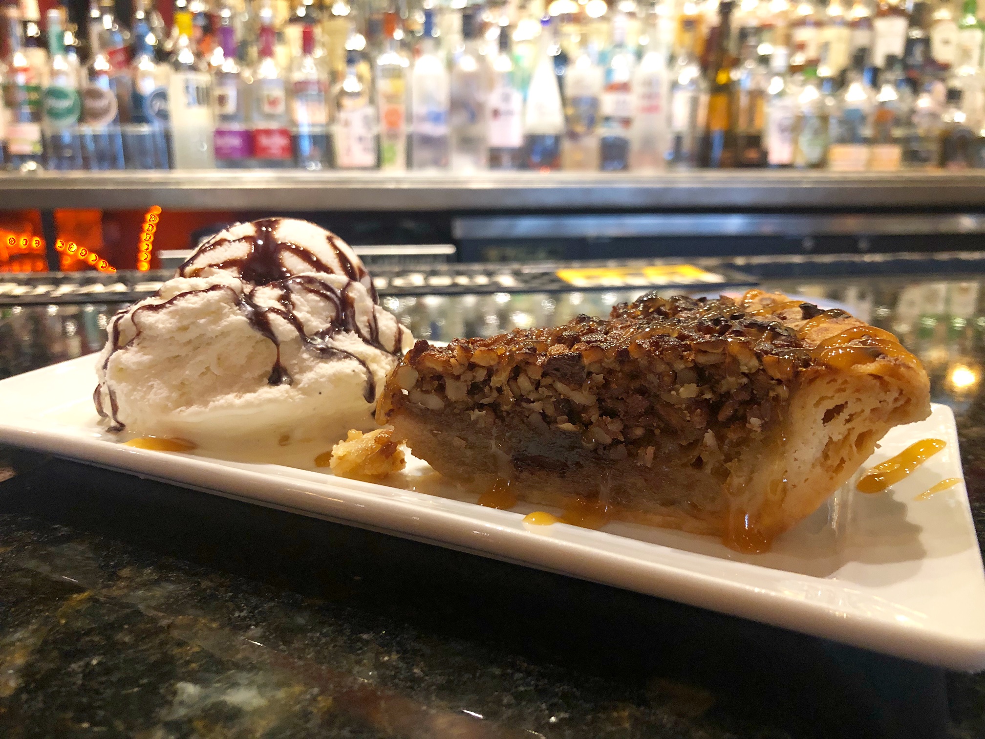 On a shiny gray bar, there is a long rectangular plate with a slice of pecan pie and a scoop of vanilla ice cream with chocolate drizzle. Photo by Alyssa Buckley.