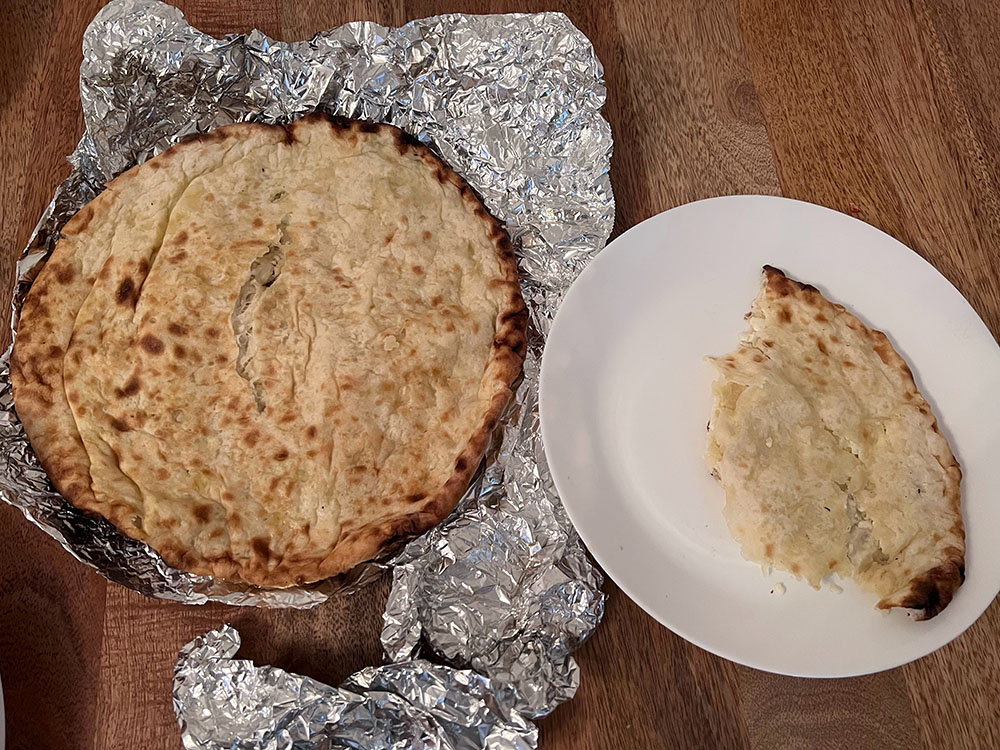 This is a photo of the paneer kulcha. On the left side of this photo is a large circe of naan break on aluminum foil on a wooden table background. On the right side is a slice of the same bread on a white plate. The naan is various shades of brown and stuffed with paneer which can faintly be seen as little chunks of white. Photo by Sara Ressing.
