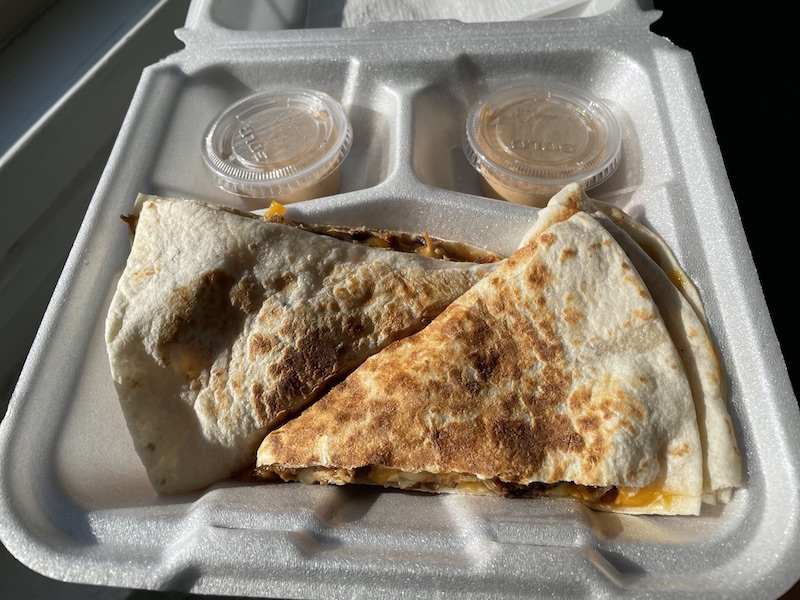 In a styrofoam box, there is a quesadilla sliced in triangles with takeout sauce in small covered plastic cups. Photo by Anthony Erlinger.