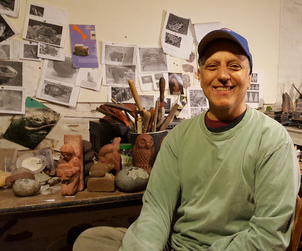 Image of Chris Berti, smiling, in front of multiple photographs and clay pieces strewn about in his colorful study. 
