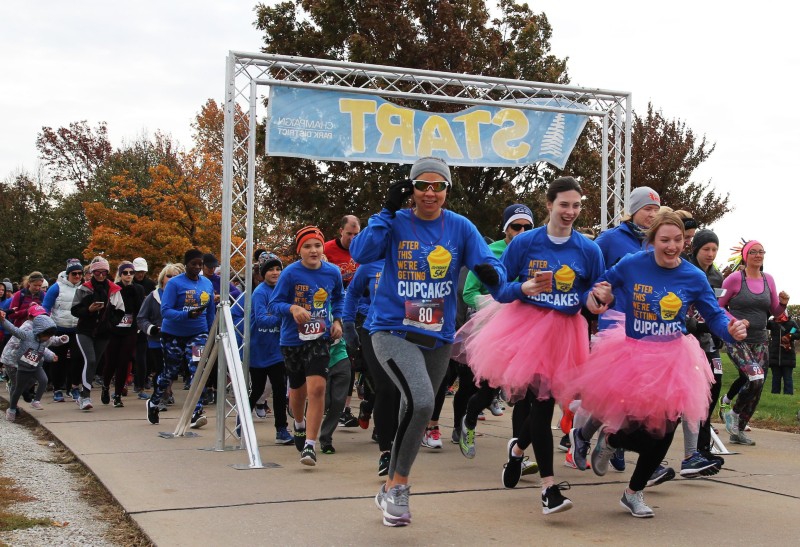 Dozens of people are running through a starting gate. They are dressed in caps and warm running clothes, and a few people have pink tutus on. Photo from Champaign-Urbana Special Recreation Facebook page. 