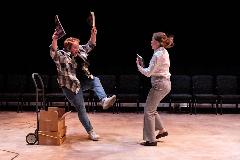 On stage, there is a white male with his arms up and one leg up in surprise in front of a white woman in dress pants and a white button up shirt. There is no audience. Photo by Bryan Heaton.