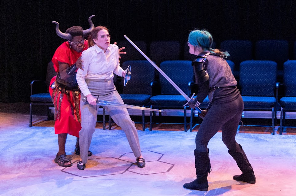 On stage, there are three characters. One character is a Black male in all red hiding behind a fighting white woman. There is a third character, a woman with bright teal hair in a ponyail holding a sword. Photo by Bryan Heaton.