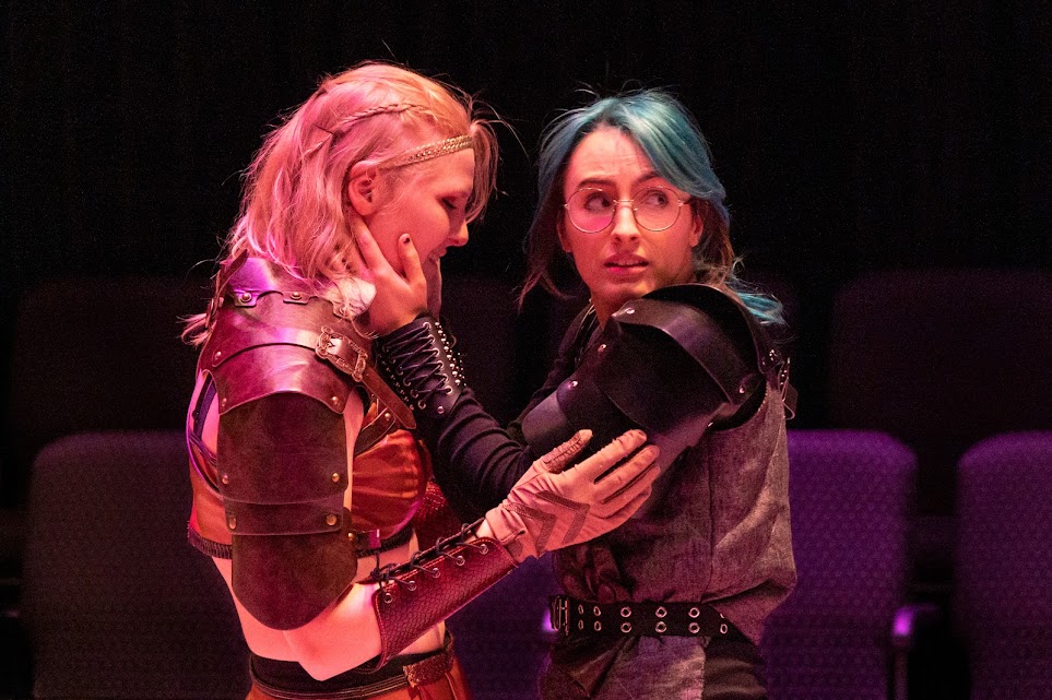 On a dark stage, two women embrace. On the left, a blonde warrior in brown holds the arms of Tilly. Tilly has teal hair and is holding the face of the other woman while looking off to the right with fear. Photo by Bryan Heaton.