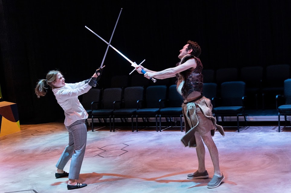 Two actors engage in swordfighting onstage. On the left, there is a white woman in work clothes fighting swords crossed a white male in basic brown warrior costume. Photo by Bryan Heaton.