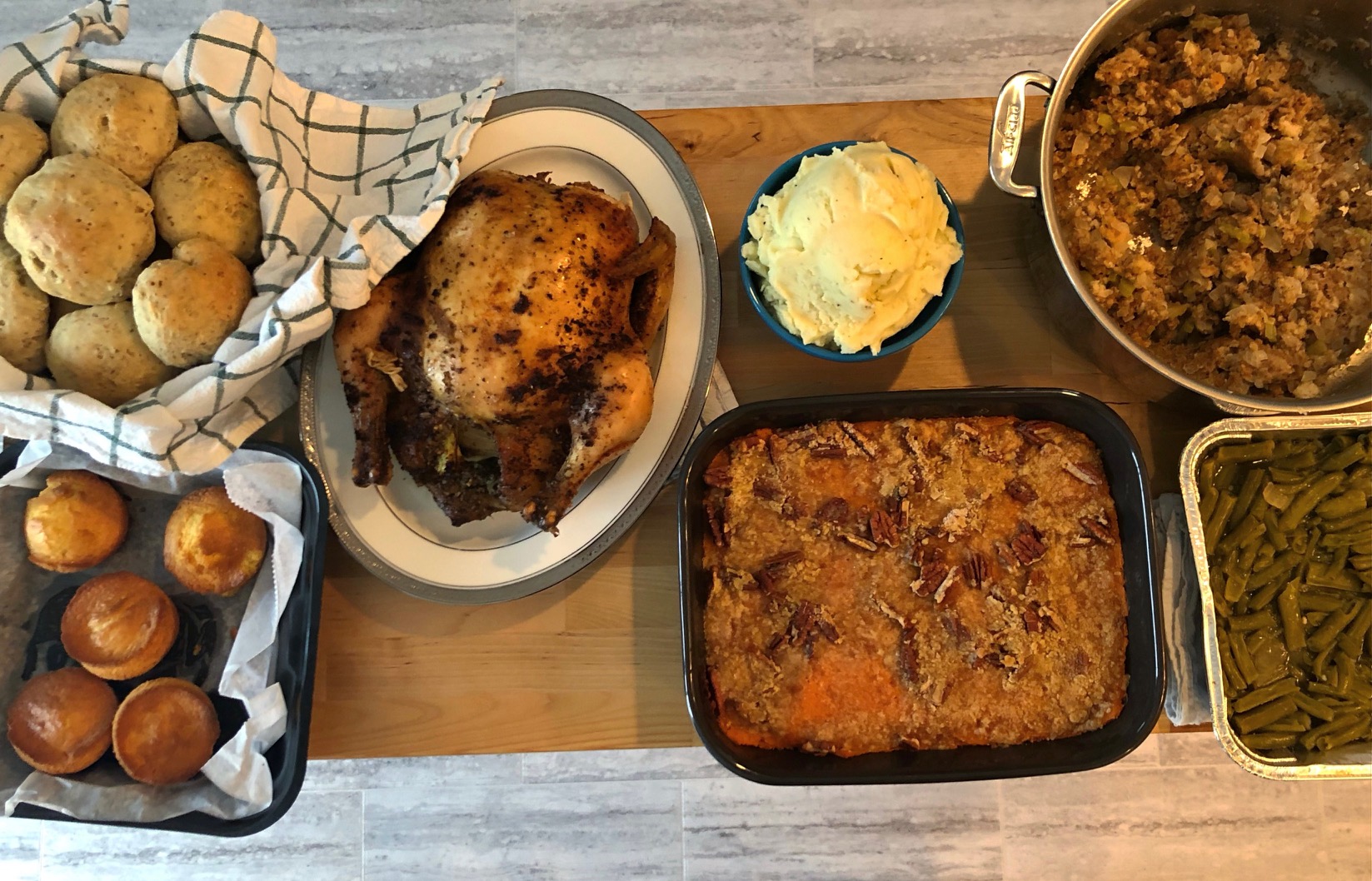 An overhead photo shows the author's Thanksgiving spread: two baskets of rolls, a whole chicken, a bowl of mashed potatoes, a tin pan of green beans, a metal pot of stuffing, and a black casserole dish with sweet potato casserole. Photo by Alyssa Buckley.