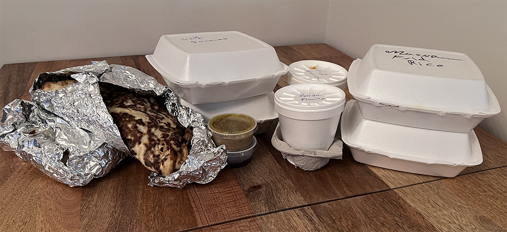 A photo of various styrofoam and foil take out containers on a wood table with white background. There is a gash of naan break bursting out of the foil. Some containers are labled with contents but text cannot be read. Photo by Sara Ressing.