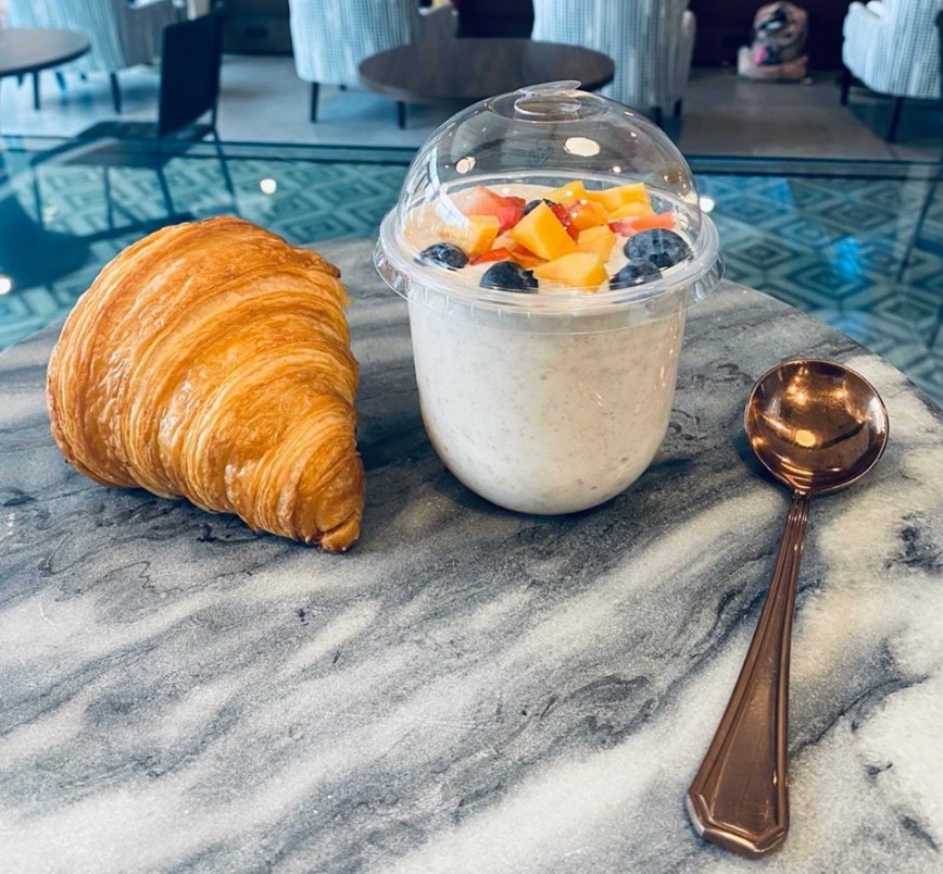 On a marble counter, there is a plain butter croissant beside a glass jar of overnight oats and a silver spoon. Photo by Dai Shi.