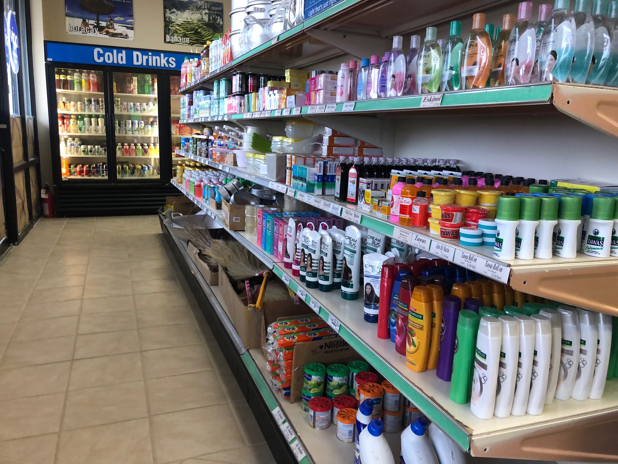 Inside the sun-soaked store, there is a front aisle of personal hygiene and cleaning items plus bottles of drinks. At the end of the aisle, there is a cooler with drinks and a blue sign that reads 