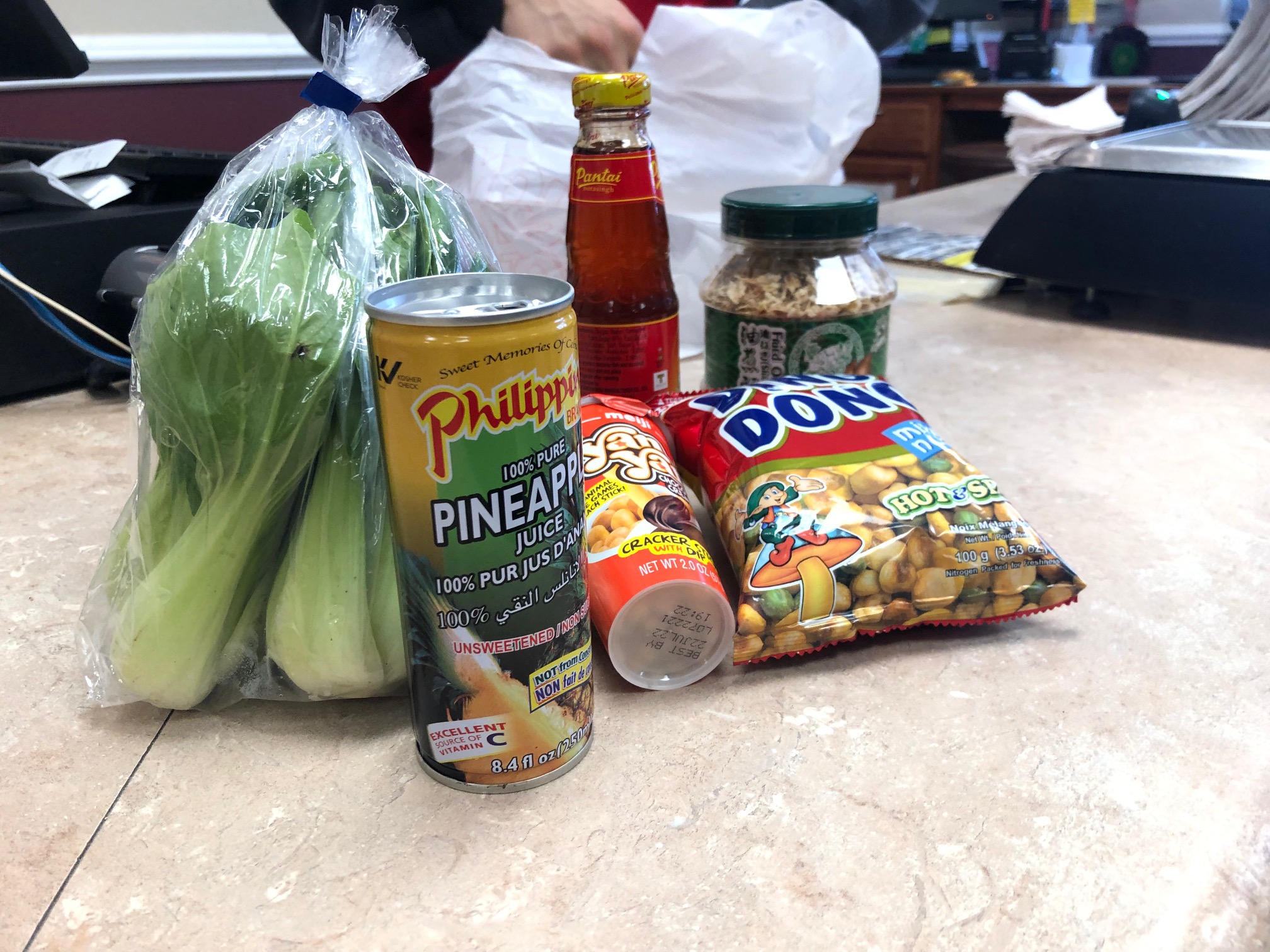 The author's purchase sits at the check out: a plastic bag of Shanghai choy, a can of pineapple juice, a jar of sauce, a cup of chocolate treats, a bag of spicy nuts, and a jar with green lid full of fried onions. Photo by Alyssa Buckley.