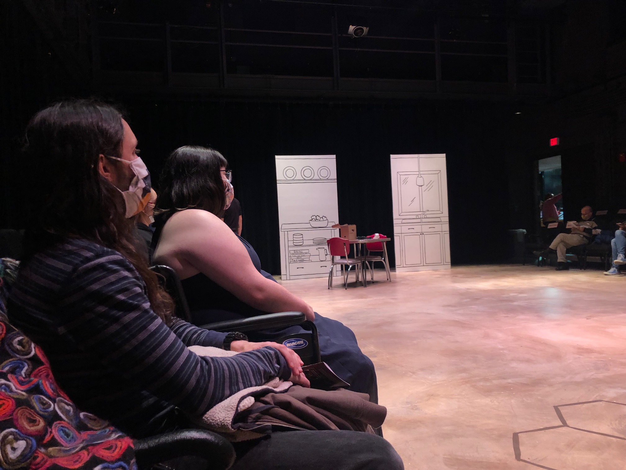 From the author's perspective in the audience, there is a set in the background of two tall white panels and a table with two chairs. Two audience members to the left of the author sit facing the stage. Photo by Alyssa Buckley.