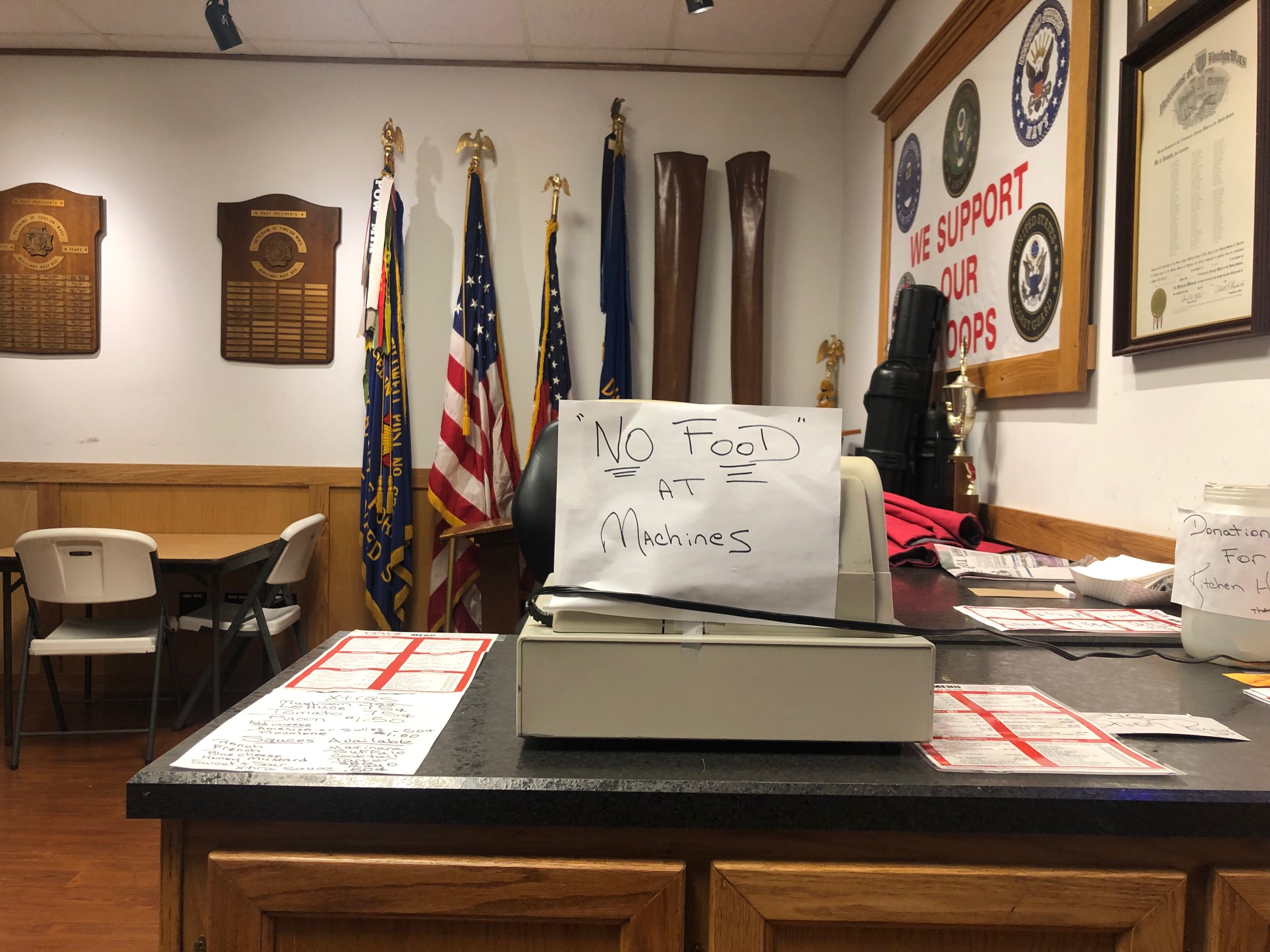 On a wooden bar, there is a cash register with a white piece of paper. The paper has a handwritten note: No Food at the Machines. Photo by Alyssa Buckley.