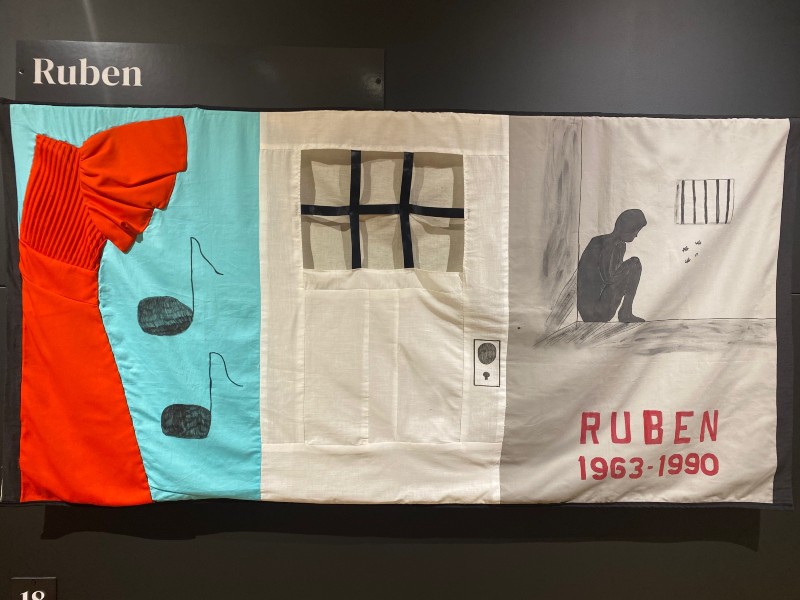A three-paneled rectangular quilt piece hanging on a black wall. One panel has musical notes and a red dress, one has a white door with black bars on the windows, and the other shows the silhouette of a man sitting on the floor of a jail cell, tally marks are scratched on the wall. The name Ruben is in white letters above the piece. Photo by Julie McClure.