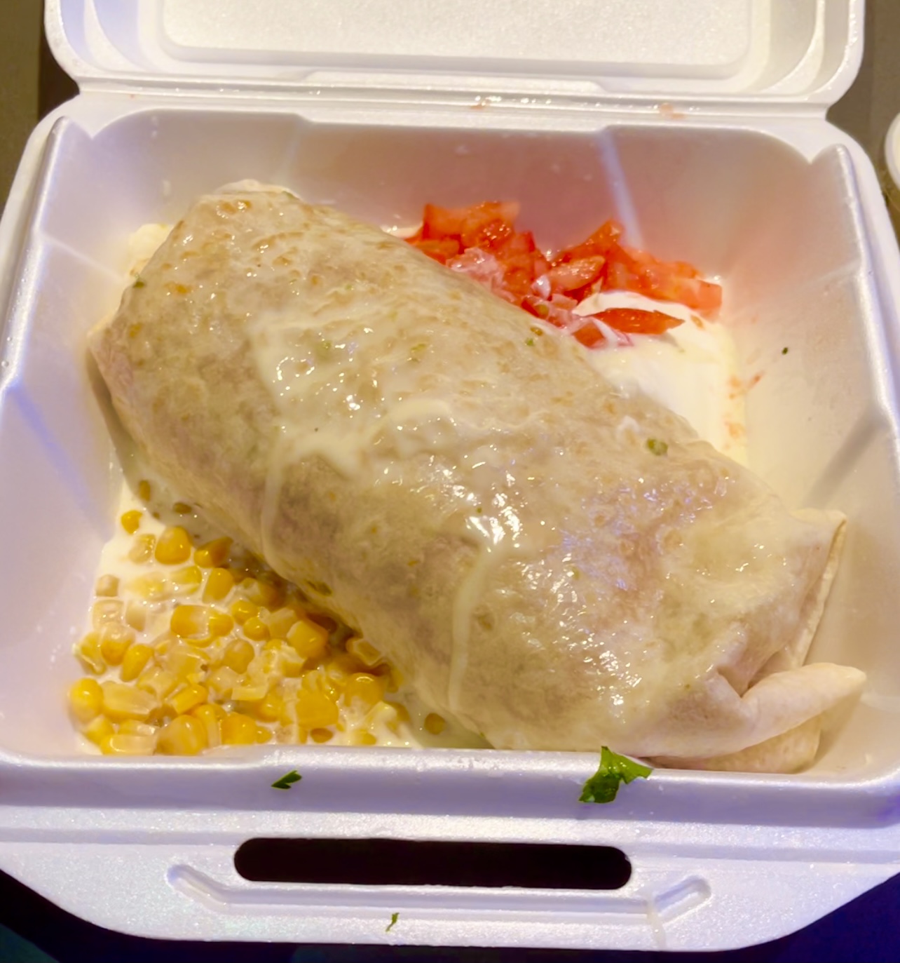 In a white styrofoam container, there is a huge burrito diagonally covering the whole container with a scoop of corn kernels and diced tomatoes on either side. Photo by Stephanie Wheatley.