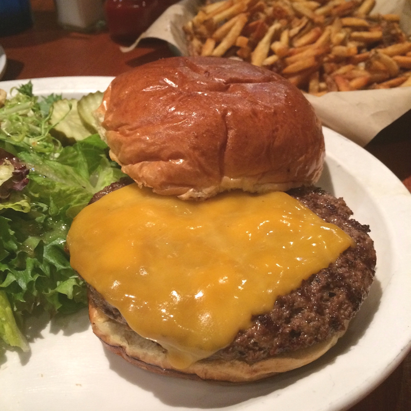 An open faced burger with melted cheddar cheese on a white plate. There is a green salad on the plate and a basket of thin cut fries in the background. Photo by Jessica Hammie.