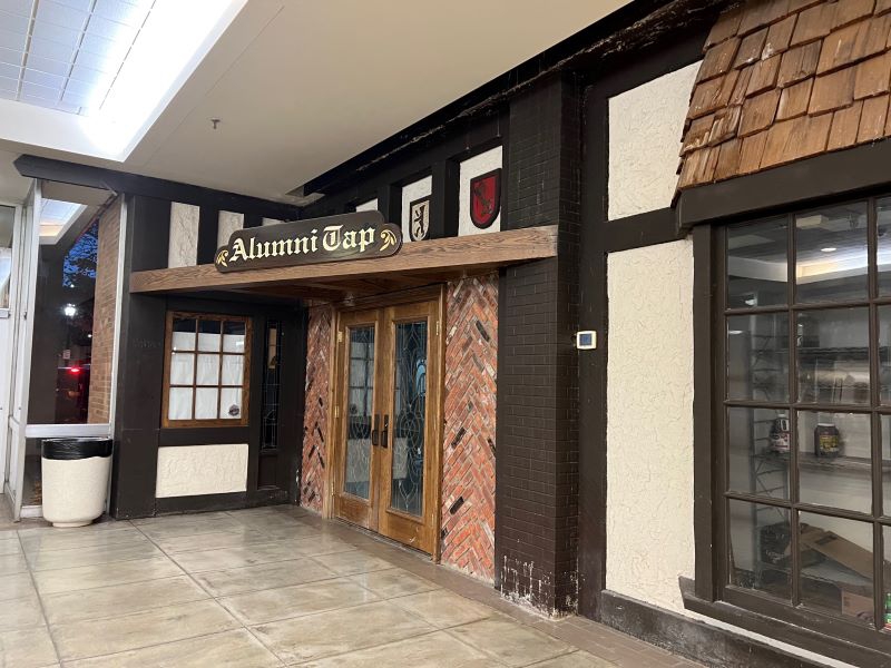 Glass doors framed in wood are flanked by brick wall. A wooden sign with Alumni Tap in stylized lettering hangs over the entrance. Photo by Julie McClure.