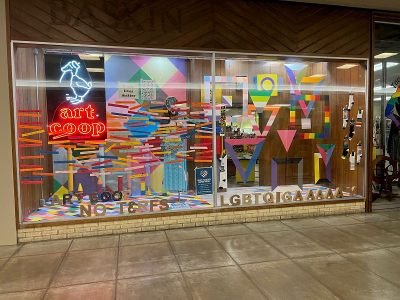 A large window with a neon sign that says art coop in red, and there is a duck with a hat in white above the words. There are colorful papers cut into shapes that reflect the colors of various LGBTQ+ flags. No Terfs and LGBTQIGAAAAAAAAAY is spelled out across the bottom. Photo by Julie McClure.