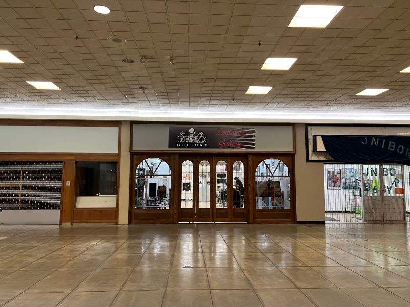 Looking across a portion of the mall, there is a storefront with several long narrow windows that are rounded at the top. Above them hangs a black sign that says Beard Culture in white, and Barbershop, Gallery in red. Photo by Julie McClure.