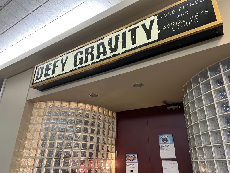 A sign with Defy Gravity in black block letters hangs over an entrance with a maroon door and glass tiled walls on either side. Photo by Julie McClure.