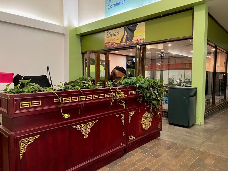 A large ornate planter with dark wood and asian-inspired detailing sits in front of a restaurant entrance that is bright green. A yellow sign that says Stango in black script with a depiction of an African woman carrying a bowl on her head hangs above the entrance. Photo by Julie McClure.