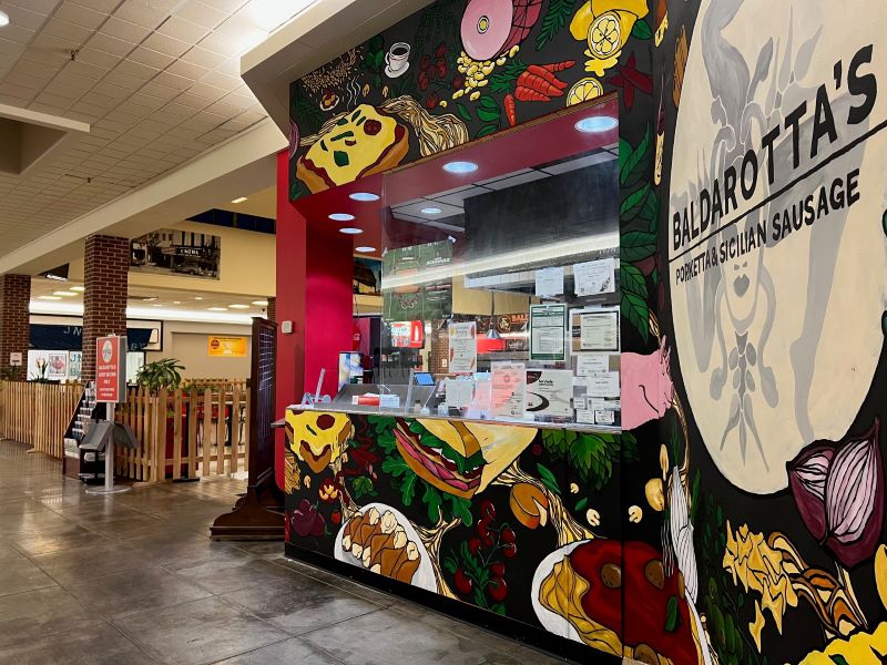 A wall with a black background painted with large cartoonish depictions of pizza, sandwiches, and other foods. A sign on the right side says Balderotta's Porketta and Sicilian Sausage. Photo by Julie McClure.